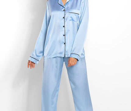 Female model standing barefoot against a white background wearing a light blue satin loose-fit button-down pyjama top with black piped trim and matching straight-leg pyjama trousers