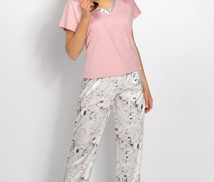 A female model stands barefoot against a plain white wall wearing a loose-fitting pink cotton V-neck short-sleeve top and light grey satin wide-leg floral print trousers