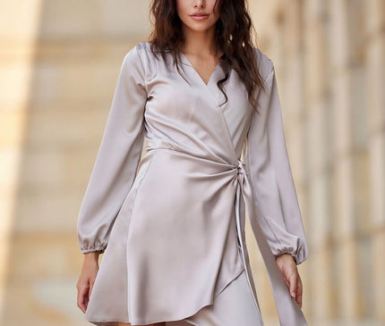 Female model poses in a light grey satin wrap mini dress with long sleeves and tie waist against a beige stone colonnade background