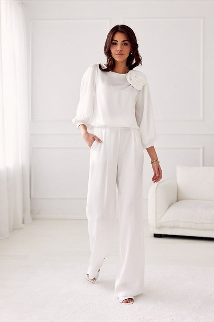 A female model standing in a white paneled room with a white sofa wears a white chiffon long-sleeve blouse with a floral shoulder detail and matching wide-leg crepe trousers while stepping forward with one hand in her pocket