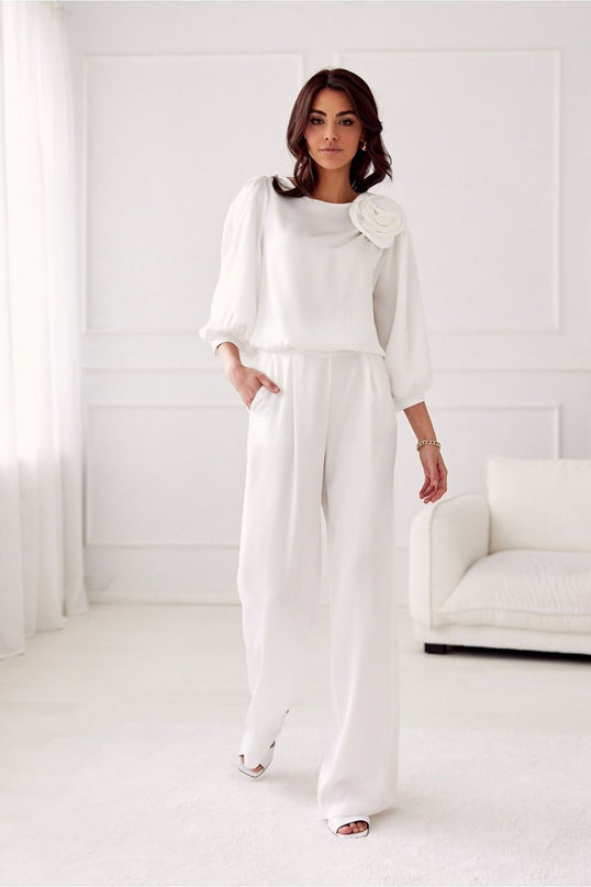A female model standing in a white paneled room with a white sofa wears a white chiffon long-sleeve blouse with a floral shoulder detail and matching wide-leg crepe trousers while stepping forward with one hand in her pocket