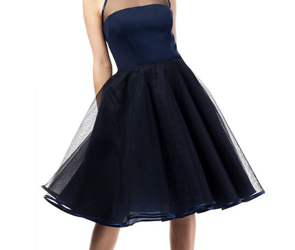 Female model in a navy blue spaghetti strap mesh yoke fit and flare tulle midi dress standing with legs apart against a plain white background