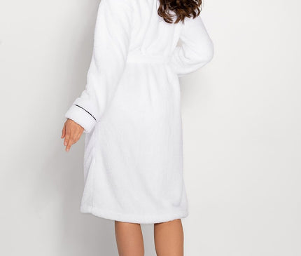 Female model in a white knee-length terry cloth bathrobe with black piping seen from the back with her head turned to the side standing barefoot against a plain light grey wall and floor