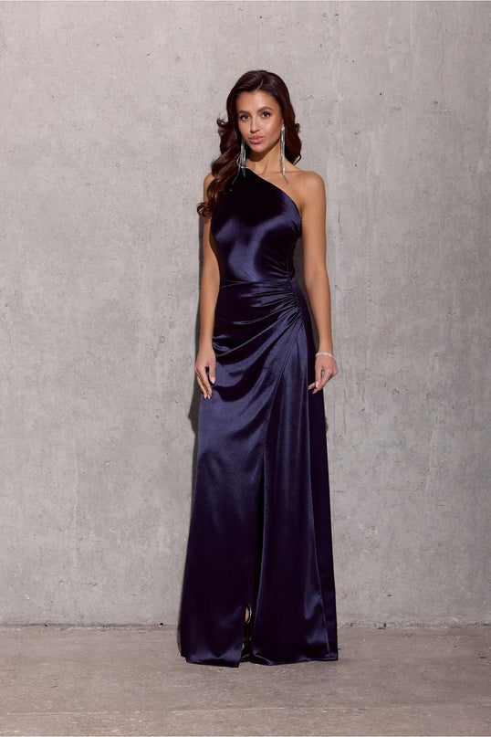 A female model stands straight against a bare concrete wall wearing a navy satin one-shoulder floor-length gown with ruched waist detailing