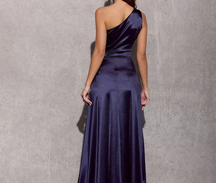 Model wearing a navy blue satin one-shoulder gown with a fitted waist and flared skirt poses with her back to camera against a bare concrete wall and floor