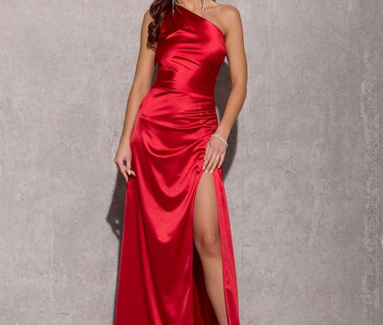 A model wearing a red satin one-shoulder floor-length evening gown with a thigh-high slit stands against a bare concrete wall and floor