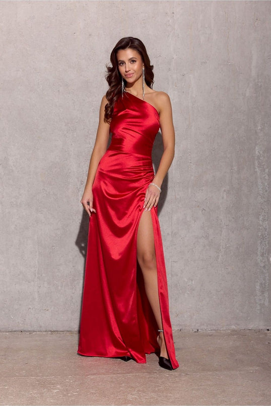 A model wearing a red satin one-shoulder floor-length evening gown with a thigh-high slit stands against a bare concrete wall and floor
