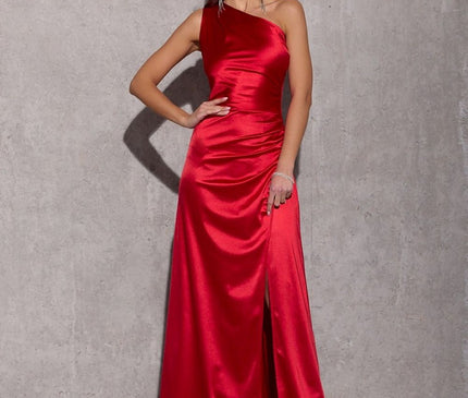 Model wearing a red satin one-shoulder floor-length evening gown with a thigh-high side slit standing against a concrete wall