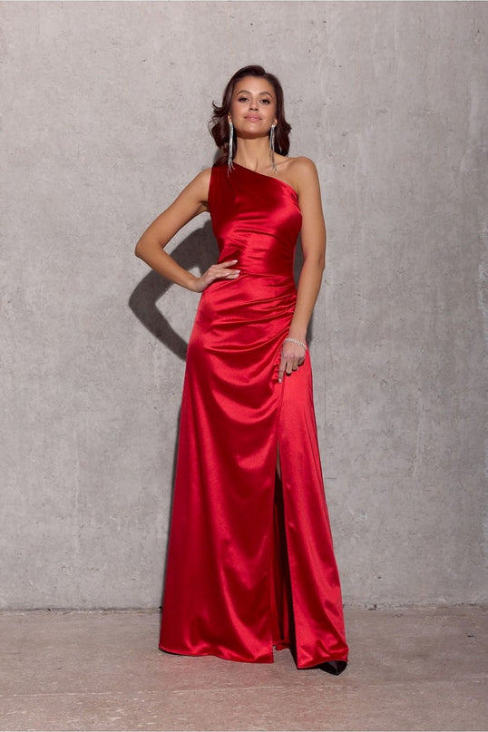 Model wearing a red satin one-shoulder floor-length evening gown with a thigh-high side slit standing against a concrete wall