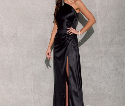 A model stands with one hand on her hip against a bare concrete wall wearing a black satin one-shoulder floor-length evening gown with a thigh-high side slit