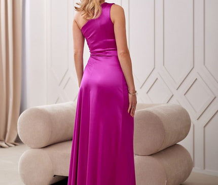 Female model in a magenta satin one shoulder maxi dress with a fitted bodice and flowing skirt poses with her back to camera in a neutral room featuring a curved beige sofa and white paneled wall