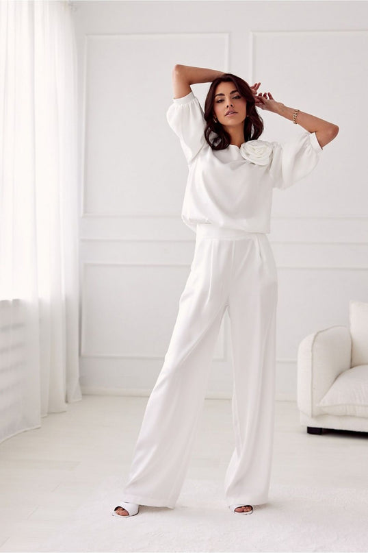 Model wearing a white satin blouse with elbow-length sleeves and a floral appliqué paired with wide-leg trousers posing in a bright white room with sheer curtains and a white upholstered sofa