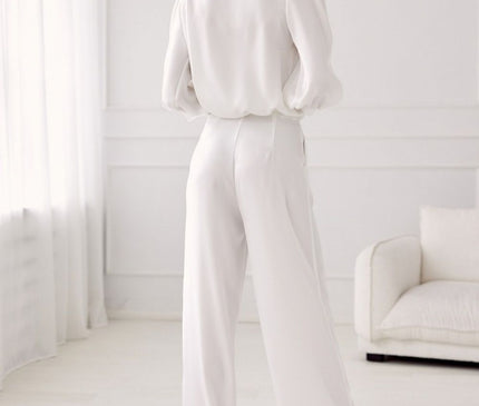 Female model wearing a white chiffon puff-sleeve blouse and wide-leg crepe trousers poses with her back to camera in a minimalist white room with sheer curtains and a white sofa