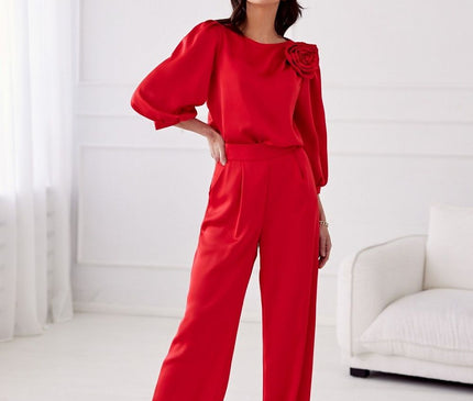 A female model stands one hand on hip in a white minimalist room wearing red satin wide-leg trousers and a matching long-sleeve blouse with a rose shoulder embellishment