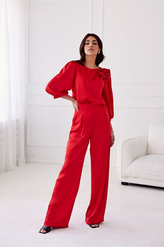A female model stands one hand on hip in a white minimalist room wearing red satin wide-leg trousers and a matching long-sleeve blouse with a rose shoulder embellishment
