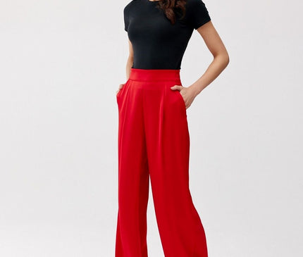 Female model in a plain white studio poses with hands in pockets wearing fluid high-waisted red wide-leg trousers and a black fitted short-sleeve top looking to the side