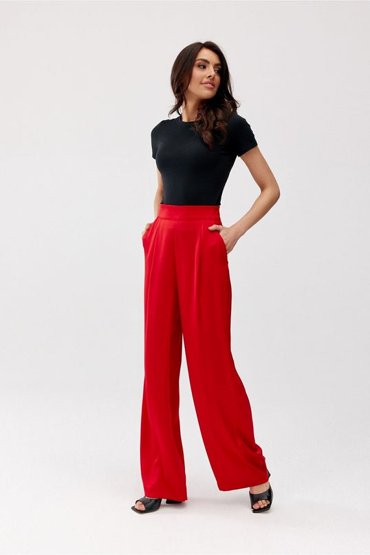 Female model in a plain white studio poses with hands in pockets wearing fluid high-waisted red wide-leg trousers and a black fitted short-sleeve top looking to the side