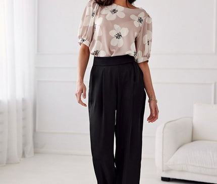 A model stands in a white room wearing a beige silky floral puff-sleeve blouse and black high-waisted wide-leg trousers posed casually beside a cream sofa against a white wall and sheer curtains
