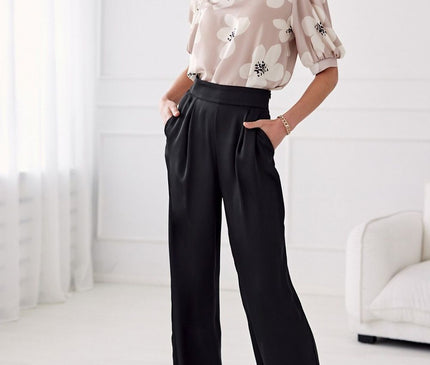 Model standing in a bright white room with sheer curtains and cream sofa wearing a beige chiffon blouse with white blossom print and puff sleeves paired with high waist wide leg black satin trousers with hands in pockets