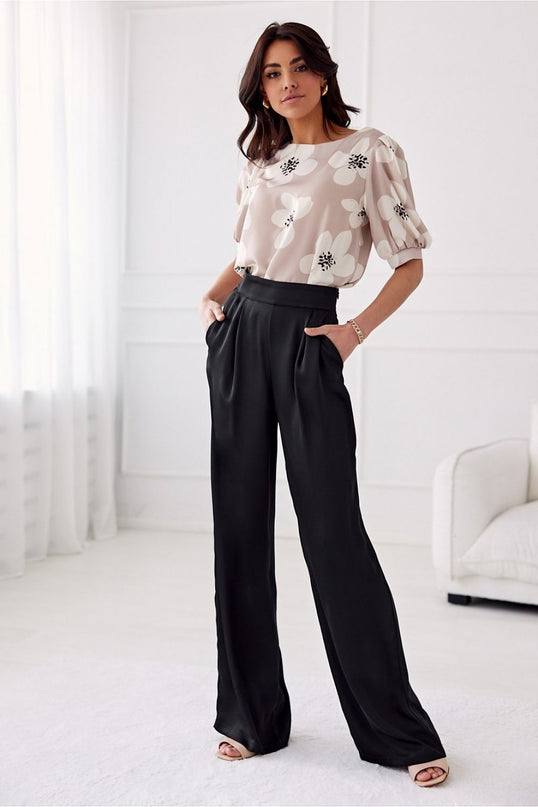 Model standing in a bright white room with sheer curtains and cream sofa wearing a beige chiffon blouse with white blossom print and puff sleeves paired with high waist wide leg black satin trousers with hands in pockets