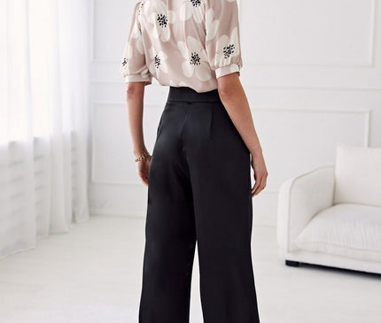 Model wearing a beige chiffon floral puff-sleeve blouse tucked into black high-waisted wide-leg trousers standing in a white-walled room with sheer curtains and a white sofa