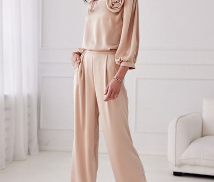 A female model stands with one hand in her pocket wearing beige satin wide-leg trousers and a matching loose-fitting long-sleeve blouse with rosette detail in a white room with moulded walls and a sofa