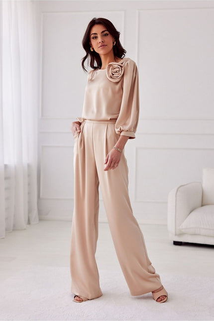 A female model stands with one hand in her pocket wearing beige satin wide-leg trousers and a matching loose-fitting long-sleeve blouse with rosette detail in a white room with moulded walls and a sofa