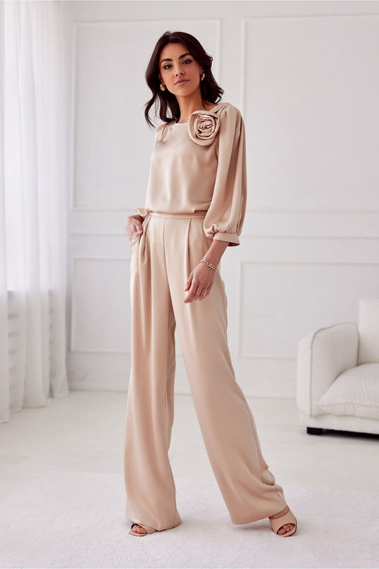A female model stands with one hand in her pocket wearing beige satin wide-leg trousers and a matching loose-fitting long-sleeve blouse with rosette detail in a white room with moulded walls and a sofa