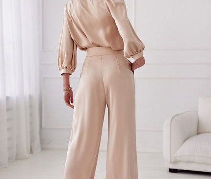 Female model wearing a beige satin balloon sleeve blouse and matching high-waisted wide-leg trousers stands facing away with one hand in pocket in a bright white room with paneled walls sheer curtains and a sofa