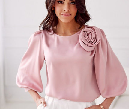 Model stands in a white minimalist room wearing a loose-fit blush pink satin blouse with voluminous balloon sleeves and a rosette shoulder detail paired with white high-waisted trousers with hands in pockets
