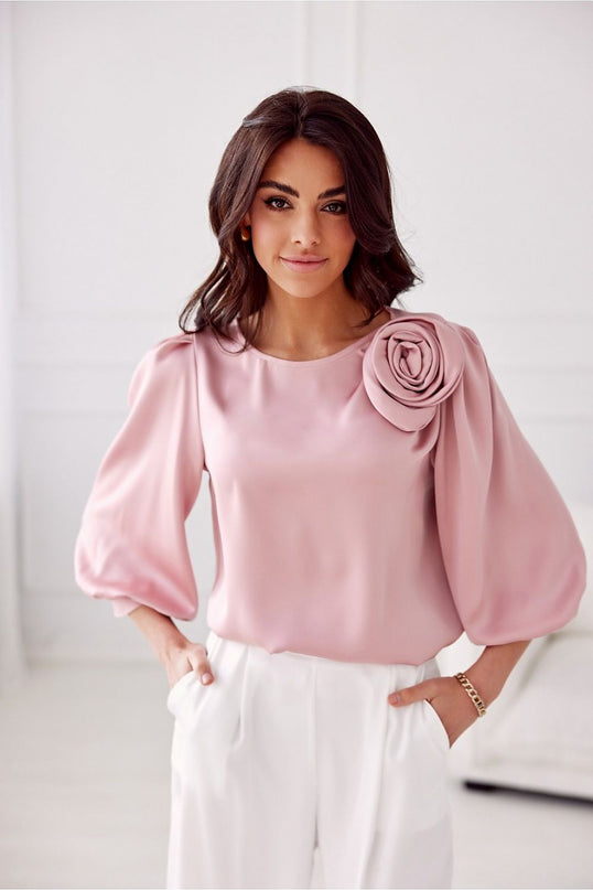 Model stands in a white minimalist room wearing a loose-fit blush pink satin blouse with voluminous balloon sleeves and a rosette shoulder detail paired with white high-waisted trousers with hands in pockets