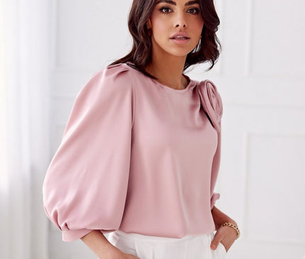 A female model stands with hands in pockets wearing a silky rose pink puff-sleeve blouse with a shoulder flower detail and white wide-leg trousers against a white panelled wall