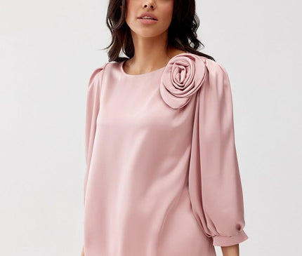 A female model stands against a plain white background wearing a loose-cut pink satin blouse with three-quarter balloon sleeves and a rose shoulder detail paired with white trousers