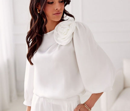 Female model in a white silk blouse with voluminous puff sleeves and rosette shoulder detail standing with one hand in pocket in a bright white room with a sofa