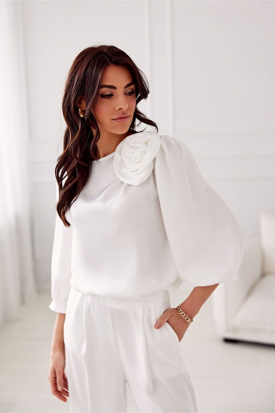 Female model in a white silk blouse with voluminous puff sleeves and rosette shoulder detail standing with one hand in pocket in a bright white room with a sofa