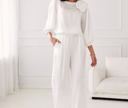 Model wearing a white chiffon blouse with puff sleeves and rose shoulder embellishment paired with wide-leg trousers standing in a bright white room with panelled walls sheer curtains and a white sofa