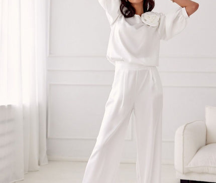 A female model wearing a white satin loose-fit blouse with a rose appliqué and matching wide-leg trousers poses with her arms raised in a minimalist white room with panelled walls and a cream sofa