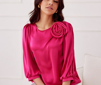 Female model seated on a white upholstered sofa against a white panelled wall wearing a hot pink satin blouse with puffed three-quarter sleeves and a rose appliqué on the shoulder
