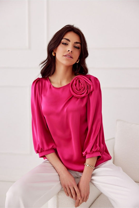 Female model seated on a white upholstered sofa against a white panelled wall wearing a hot pink satin blouse with puffed three-quarter sleeves and a rose appliqué on the shoulder
