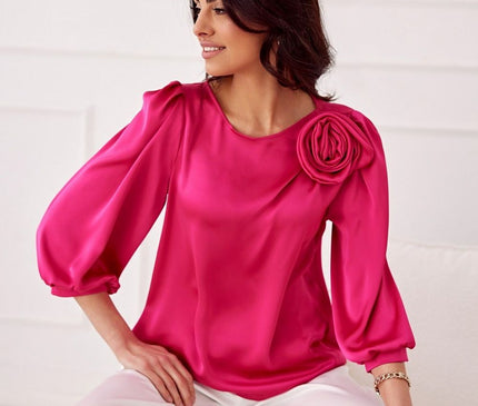 Female model seated on a white sofa in a white paneled room wearing a loose-fit fuchsia satin blouse with three-quarter puff sleeves and a rosette shoulder detail with her head turned to the side