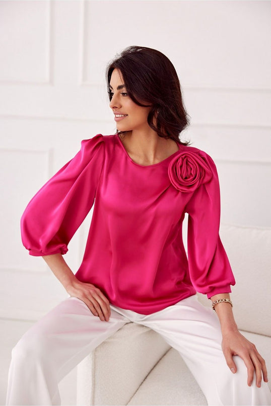 Female model seated on a white sofa in a white paneled room wearing a loose-fit fuchsia satin blouse with three-quarter puff sleeves and a rosette shoulder detail with her head turned to the side