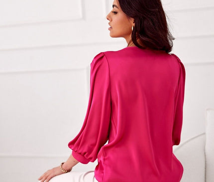 A model seated sideways on a white sofa against a panelling wall in a bright minimal room wearing a loose-fit fuchsia satin blouse with gathered cuffs
