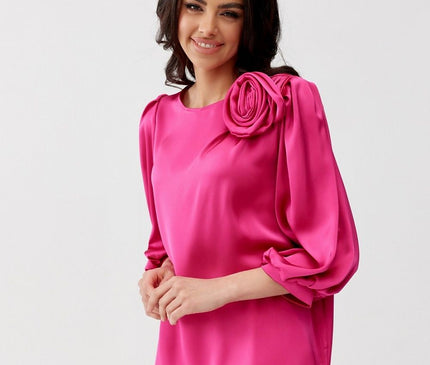 A model stands against a plain white backdrop wearing a relaxed cut fuchsia satin long-sleeve blouse with voluminous sleeves and a rosette shoulder detail paired with white trousers