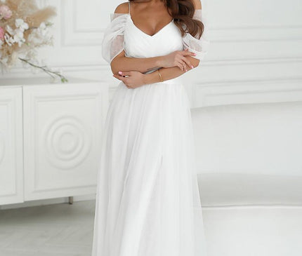 Brunette model stands with arms crossed wearing a white tulle off-shoulder maxi dress with puff sleeves in a white paneled room with a sofa and floral arrangement