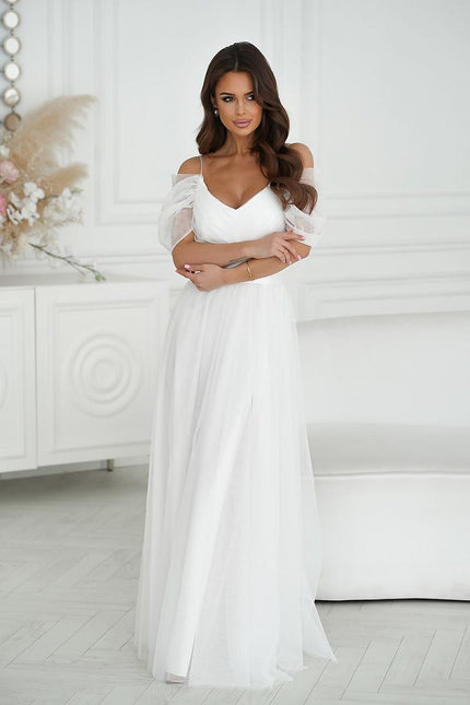 Brunette model stands with arms crossed wearing a white tulle off-shoulder maxi dress with puff sleeves in a white paneled room with a sofa and floral arrangement