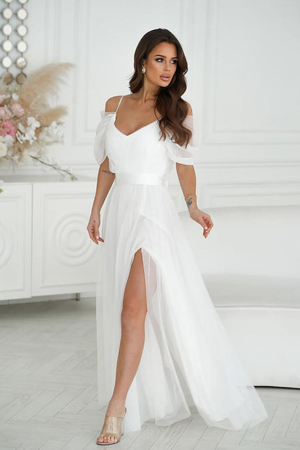 Female model wearing a white off-the-shoulder chiffon gown with a thigh-high front slit standing in a white room with paneled walls and a console table topped with pampas grass