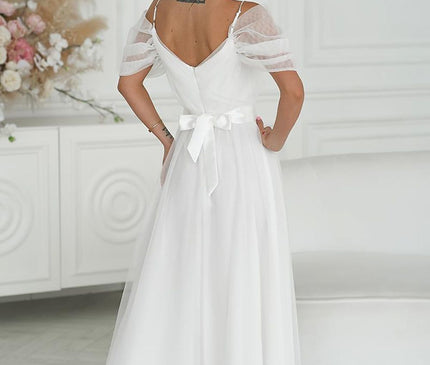 Model wearing a floor-length white chiffon off-shoulder gown with mesh puff sleeves and a satin waist tie stands facing away in a bright room with a white paneled wall a floral arrangement and round mirror discs