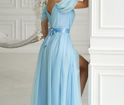 A female model leaning against a white paneled wall in a floor-length pastel blue chiffon off-the-shoulder gown with a satin waistband and thigh slit beside a floral hoop arrangement