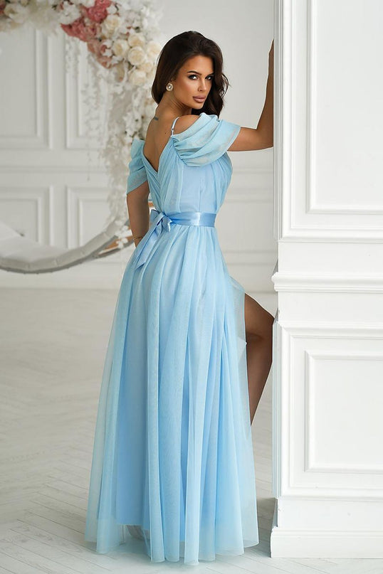 A female model leaning against a white paneled wall in a floor-length pastel blue chiffon off-the-shoulder gown with a satin waistband and thigh slit beside a floral hoop arrangement