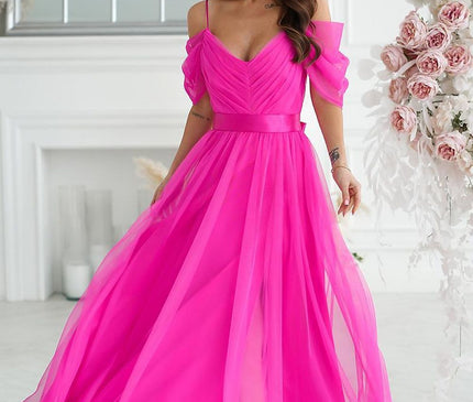 A model wearing a bright fuchsia off-the-shoulder tulle A-line gown stands holding the skirt in a white room with panelled walls a large window and a floral arrangement